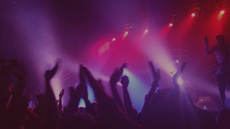 Photo taken at a concert from in the crowd. People are waving their hands in the air and purple, pink, and blue lights are flashing.